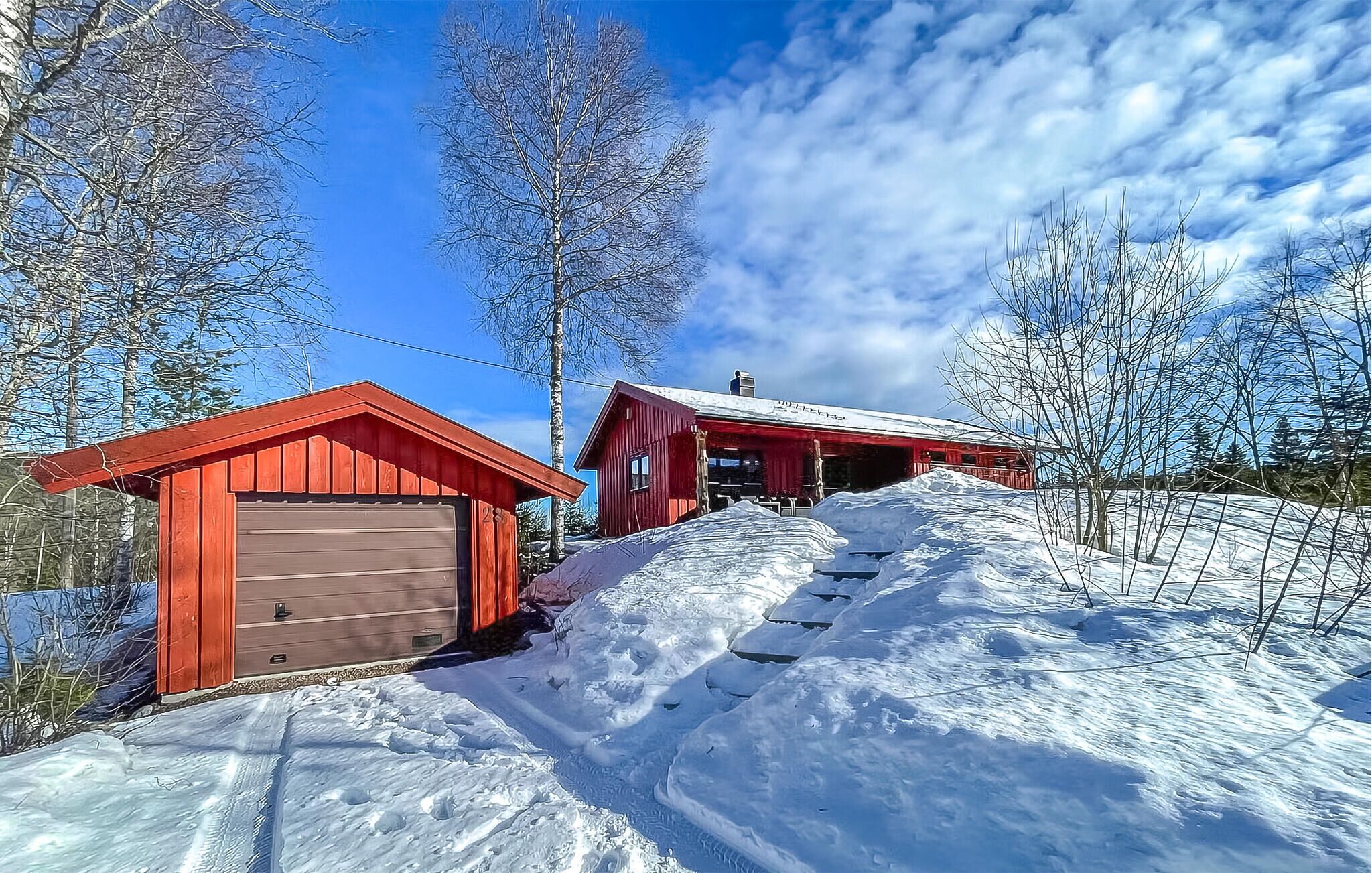 A red wooden cabin with a garage, surrounded by snow-covered ground and trees.