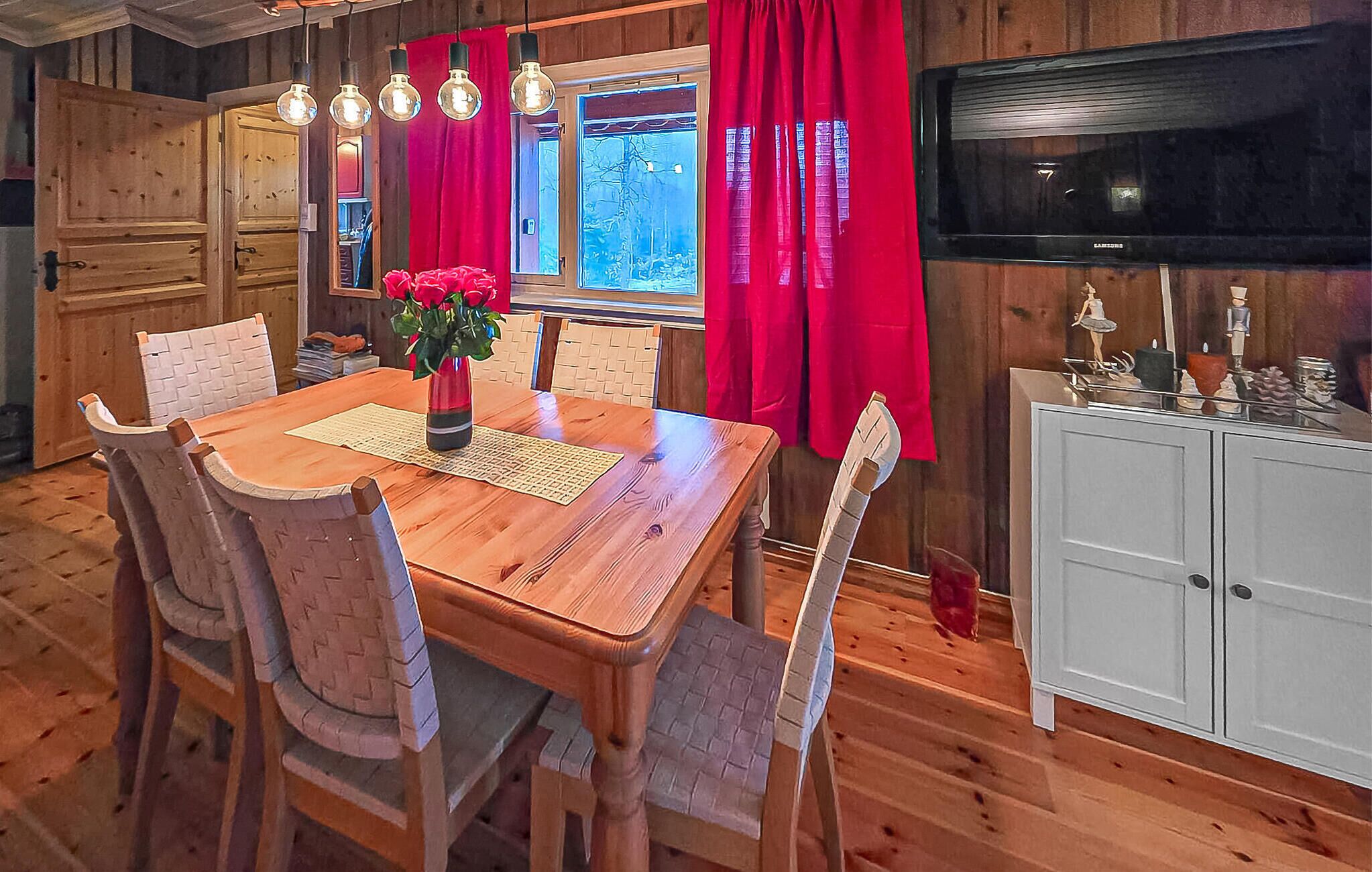 A wooden dining table with chairs, a vase with red flowers, and a television on a cabinet.