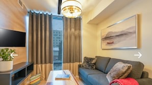Un salon moderne doté d’un téléviseur à écran plat, d’un canapé agrémenté de coussins décoratifs, d’une table basse sur laquelle repose un livre, et d’une grande fenêtre ornée de rideaux.