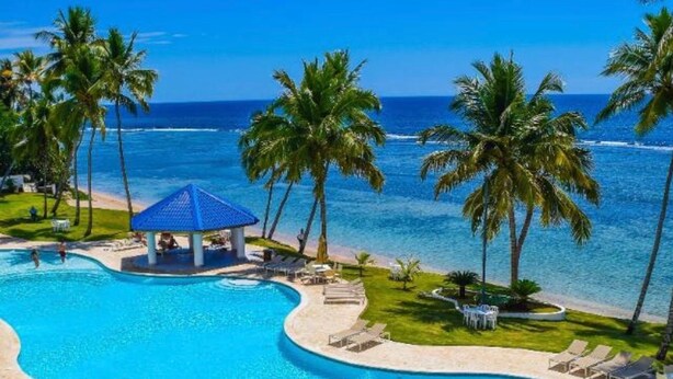 En pool med en böjd design, omgiven av palmer och med utsikt över stranden.