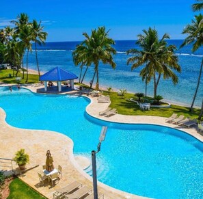 A swimming pool with a curved design, surrounded by palm trees and a beach view. - Cozy 2-bedroom Beach Front Condo with AC (Guayacanes)