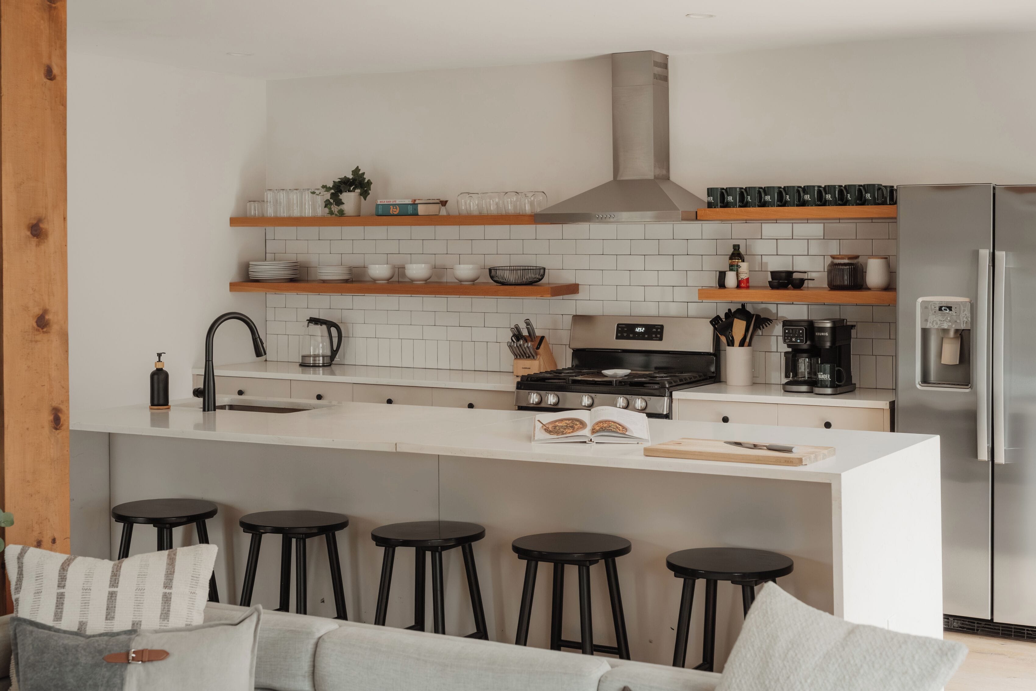 A modern kitchen with white tiles, wooden shelves, a stainless steel stove, and a built-in refrigerator.