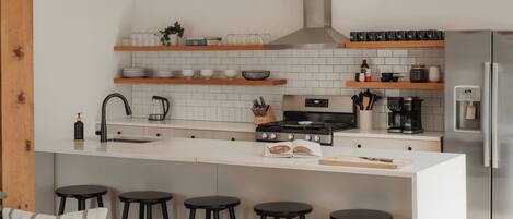 A modern kitchen with white tiles, wooden shelves, a stainless steel stove, and a built-in refrigerator.