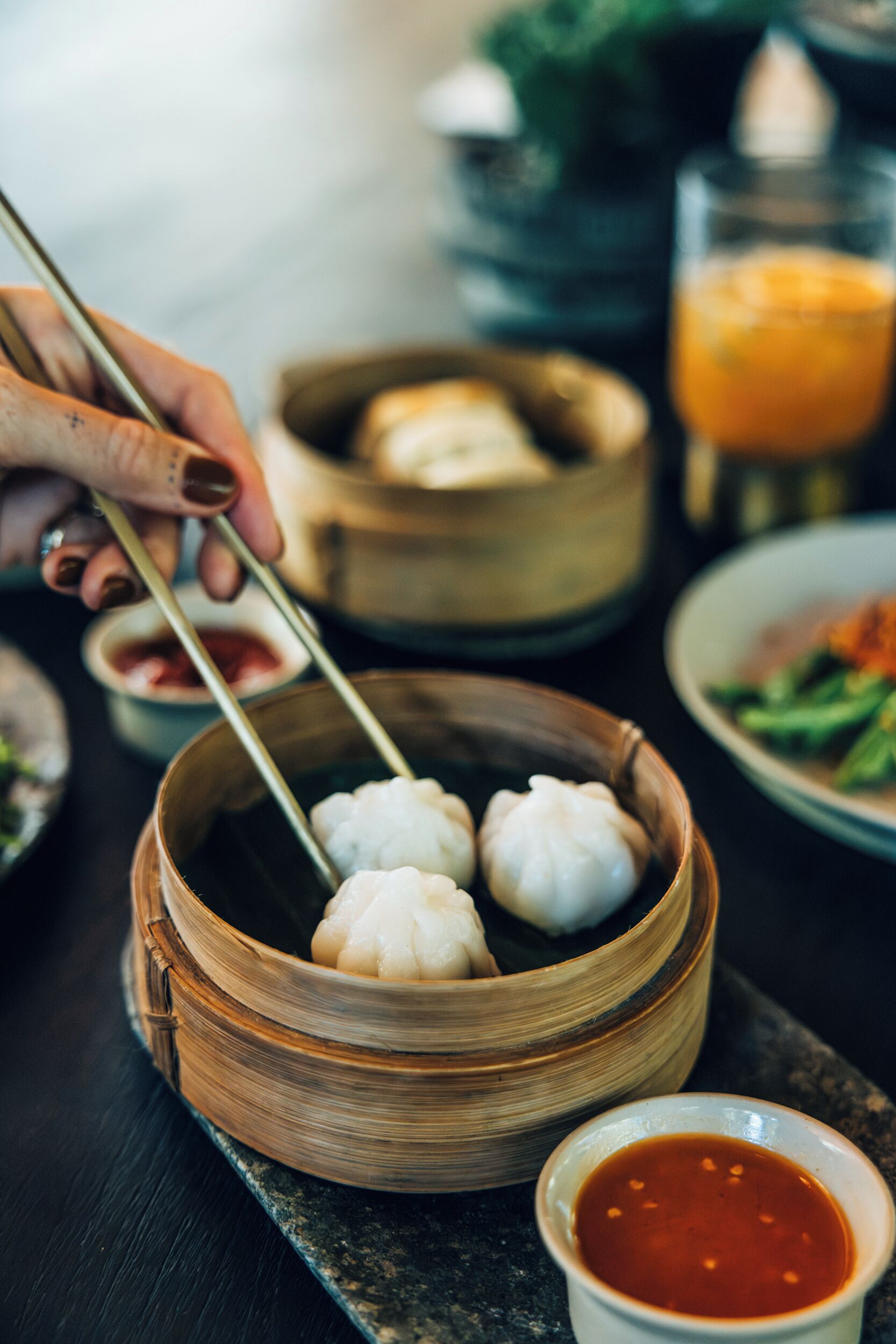 A bowl of dumplings with chopsticks, a small bowl of sauce, and a glass of orange juice on a table.