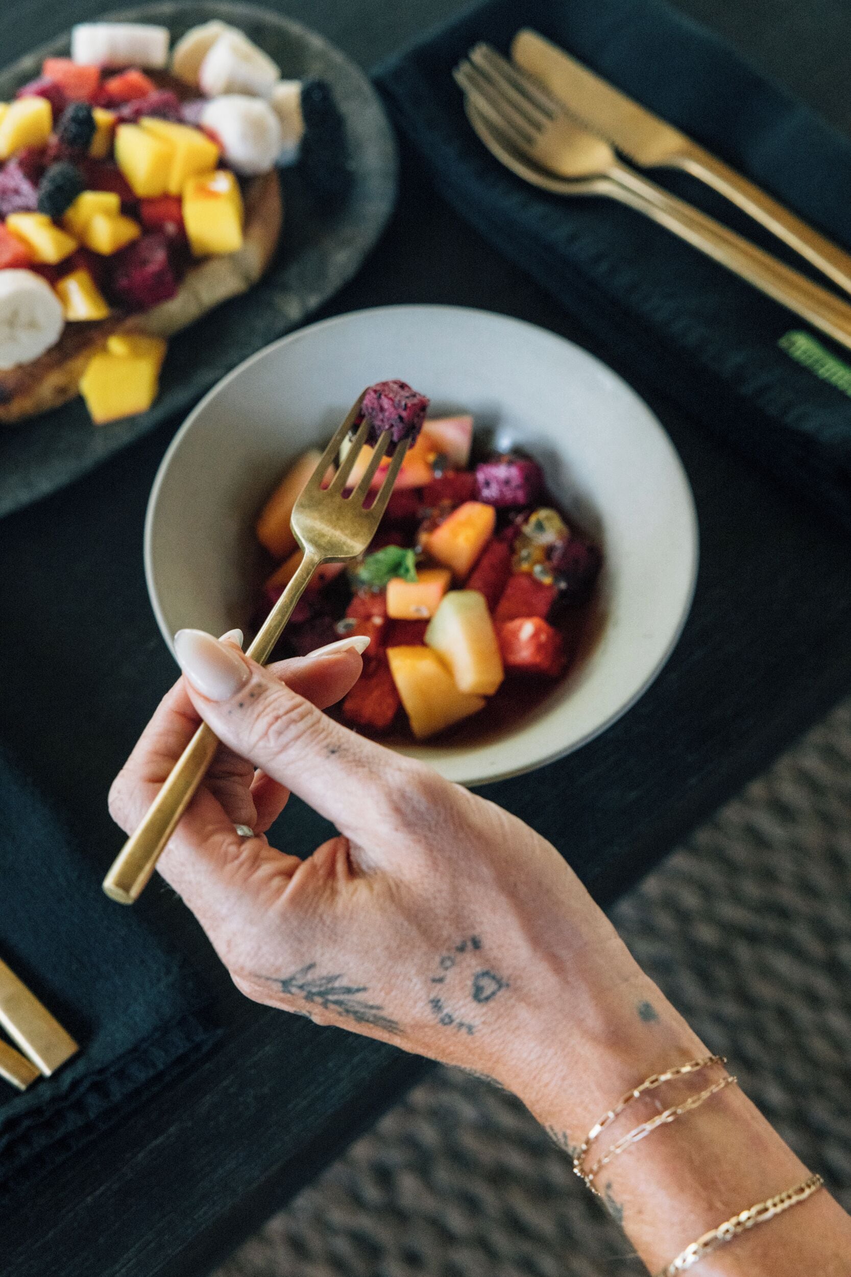 A hand holding a gold fork with a bowl of mixed fruit salad in front of it.