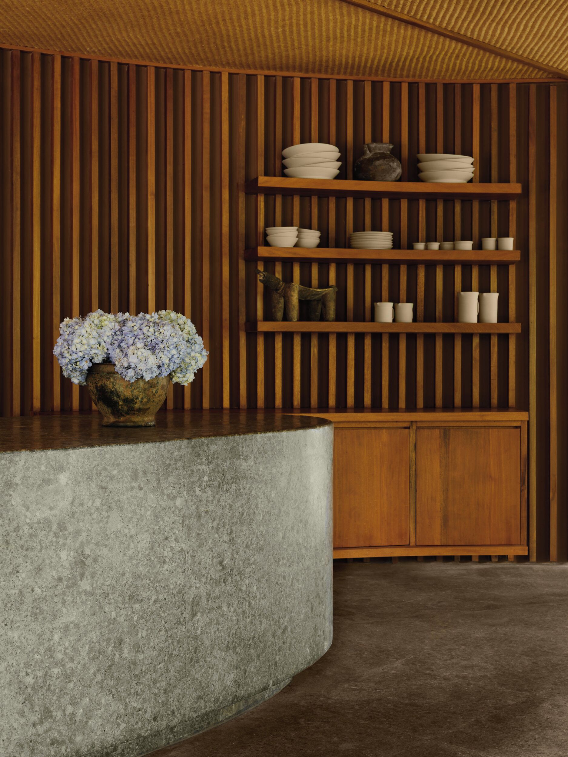 Una cocina moderna con paneles de madera, una isla de concreto y estantes con vajilla y un florero.
