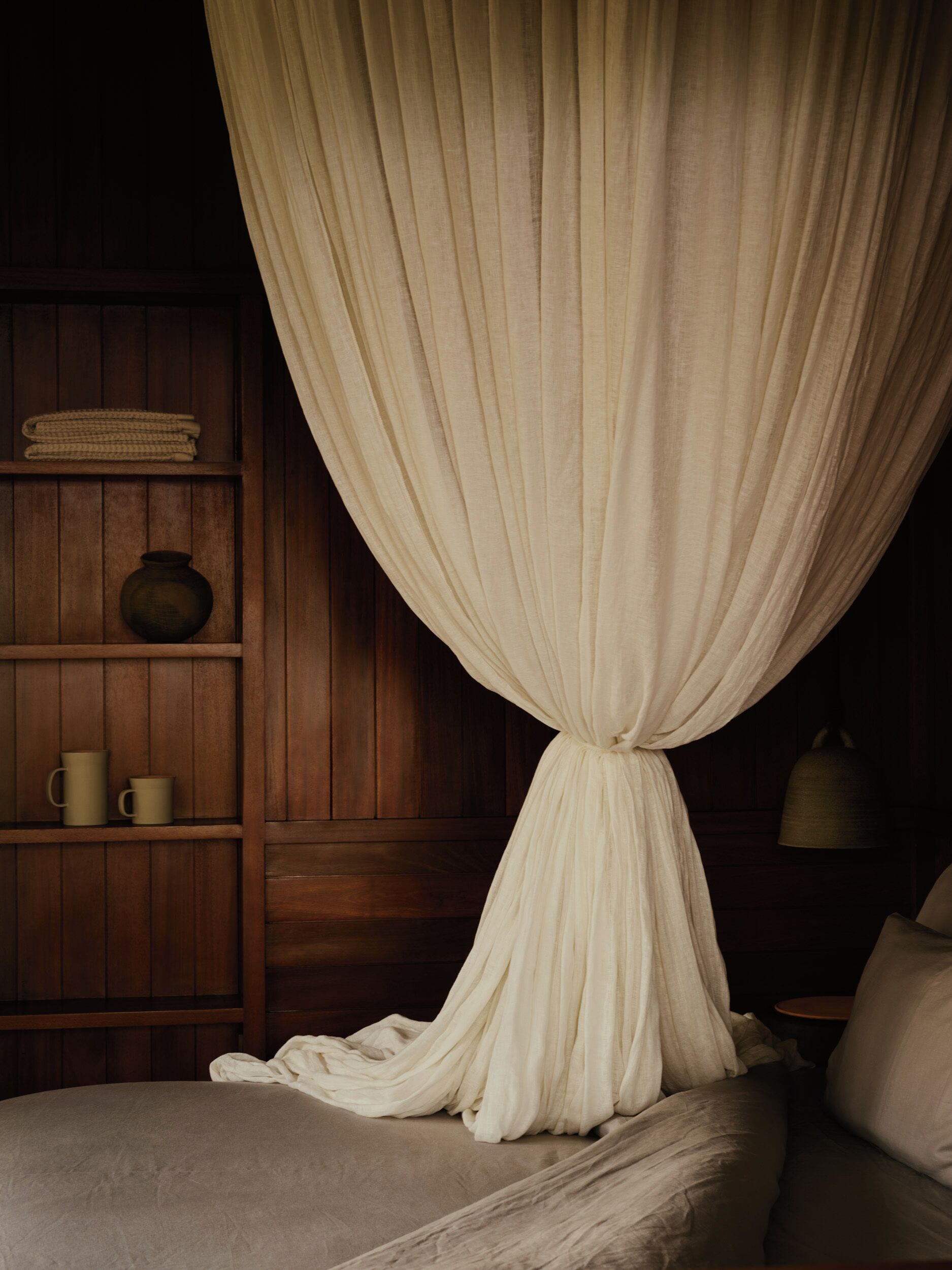 Quarto com cama, prateleira de madeira com um vaso e canecas, e um sino na parede.
