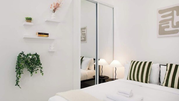 Una camera d'albergo con un letto, lampade sui comodini, uno specchio, un armadio e uno scaffale a parete con oggetti decorativi.