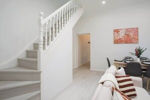 Un escalier menant à un espace repas où une table est dressée pour deux personnes, un tableau accroché au mur et un canapé agrémenté de coussins.