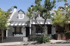 Une maison de style victorien avec un porche à l’avant, une clôture en piquets et un petit arbre dans le jardin.