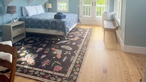 Un dormitorio con techo inclinado, una cama con colcha estampada, una mesita de noche, una silla, suelo de madera y una ventana con vistas a la vegetación.
