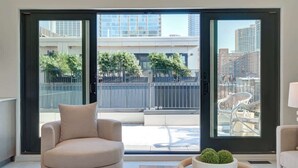 Een moderne woonkamer met een beige fauteuil, een houten salontafel met een plant en grote schuifdeuren die leiden naar een buitenbalkon met uitzicht op de stad.