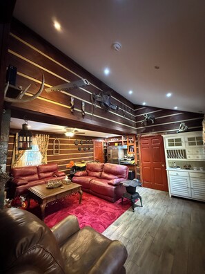 A spacious living room with a red sofa, a brown leather chair, and a wooden coffee table. A kitchen area is visible in the background with a refrigerator and cabinets. - 3-bedroom house with outdoor hot tube on 9 acres of total privacy (Boone)