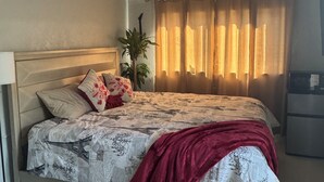 Una cama bien hecha con ropa de cama estampada, una manta roja y una planta en maceta a su lado.