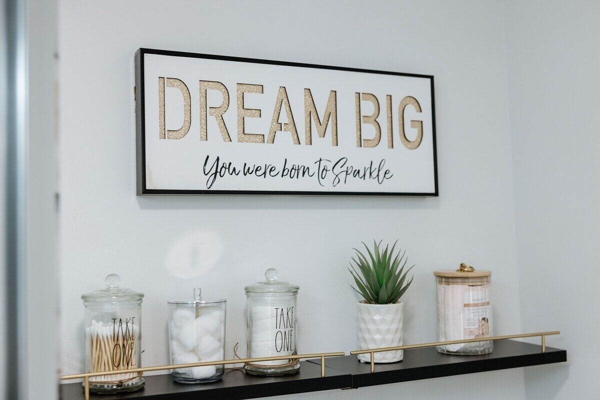 A framed motivational quote on a wall, a shelf with jars labeled "TAKE ONE," and a small potted plant.