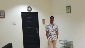 Un homme se tient dans une pièce où l’on aperçoit une porte noire, une étagère en métal et un tableau encadré accroché au mur.