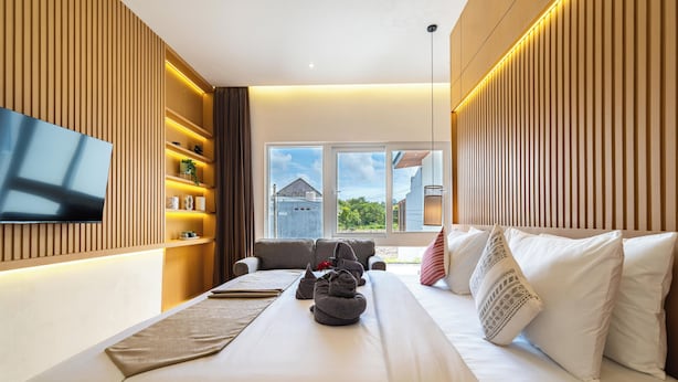 Habitación de hotel moderna con una cama grande, televisor de pantalla plana montado en la pared, repisa empotrada con objetos decorativos y vistas al exterior a través de amplios ventanales.