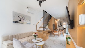 Una sala de estar moderna con un sofá beige, un sillón gris y una mesa de centro blanca. Se ve una escalera que conduce a un área de comedor con sillas de madera y una mesa.