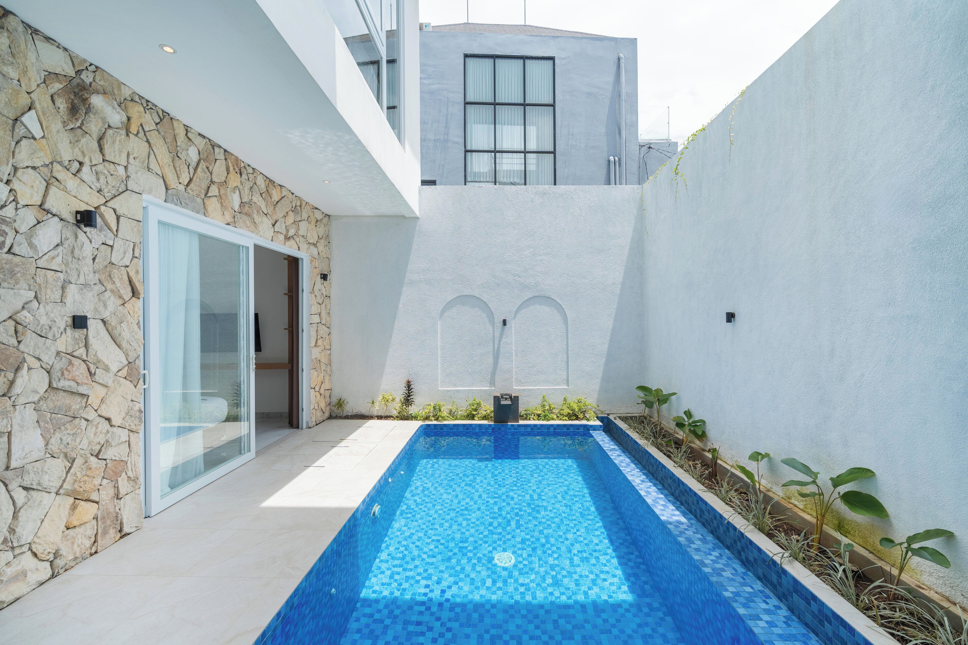 A modern outdoor swimming pool with stone and white walls, a glass door entrance, and a small garden area.