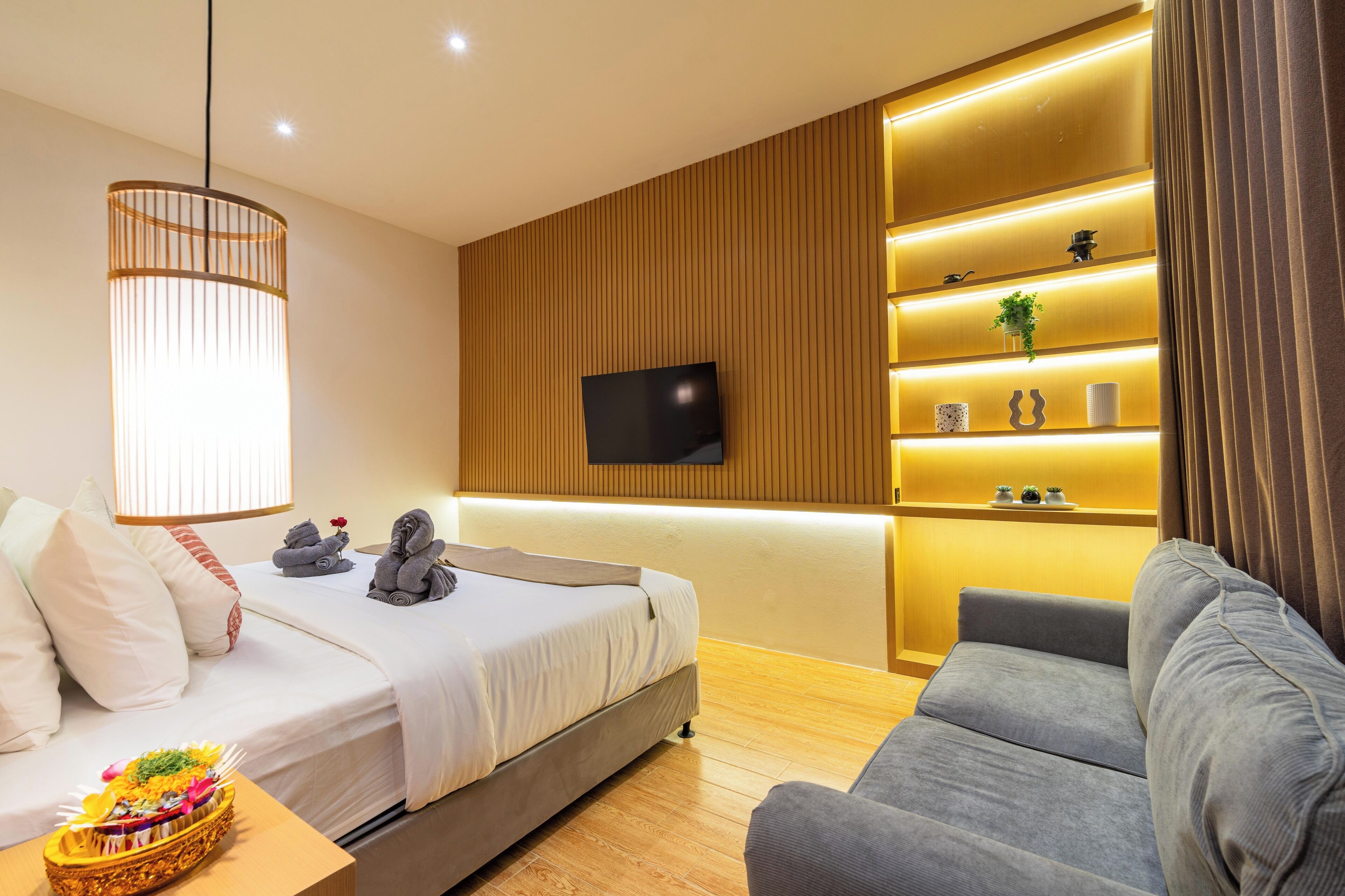 A modern hotel room with a bed, a grey sofa, a TV mounted on a wooden wall, and a shelf with decorative items.