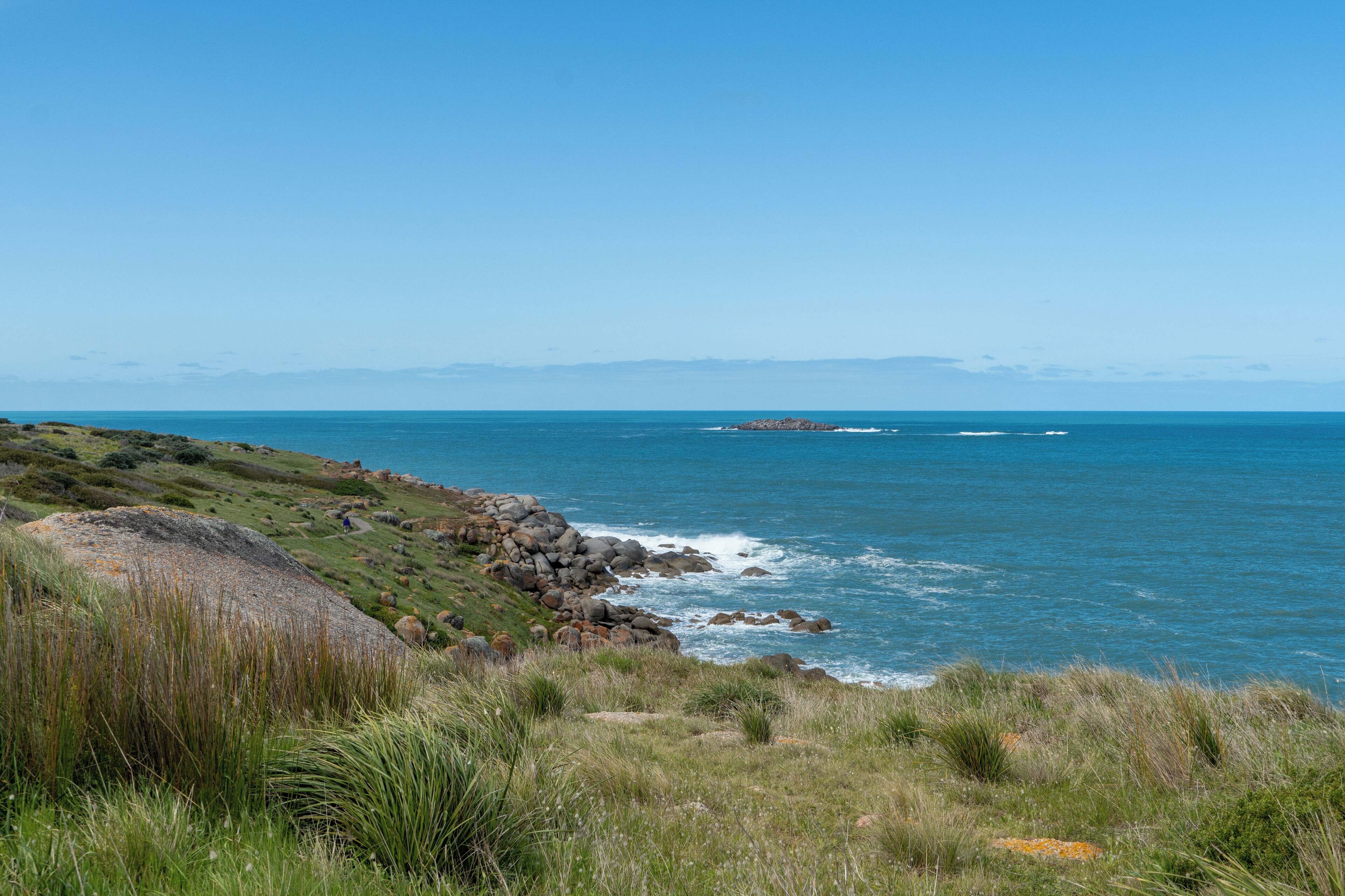 A coastal landscape with a rocky shoreline, grassy terrain, and a clear blue sky.