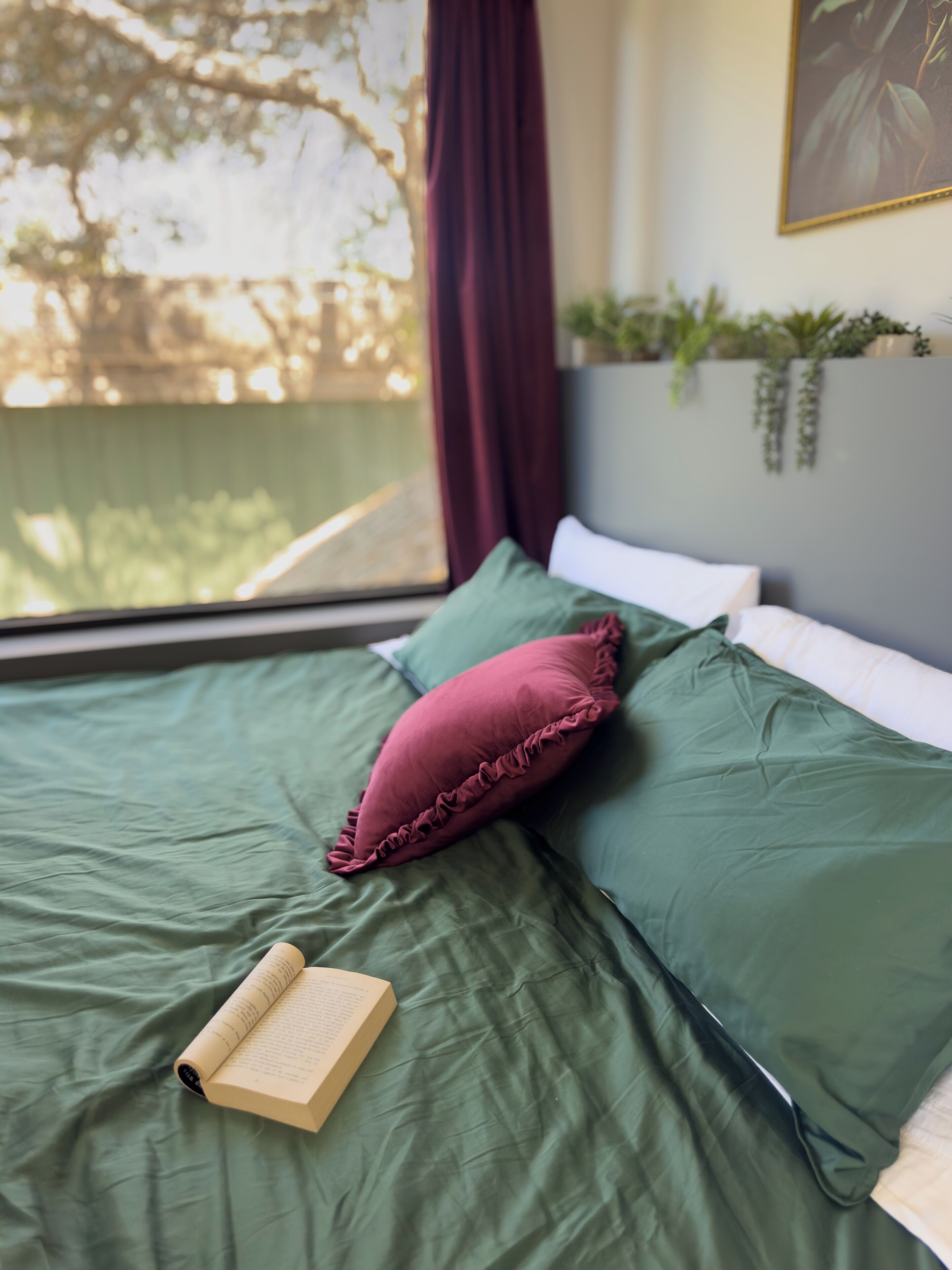 A bed with green bedding and a red pillow, a book on the bed, and a window with a view of trees.