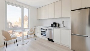 A modern kitchen with white cabinets, a stainless steel refrigerator, a coffee maker, and a dishwasher. There is a window with a view of a cityscape. - Blueground | Downtown Brooklyn, rooftop, nr Brooklyn Bridge (New York)