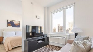 A modern hotel room with a bed, a TV, a sofa, a window with a city view, and a heater. - Blueground | Downtown Brooklyn, rooftop, nr Brooklyn Bridge (New York)