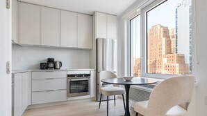 Une cuisine moderne avec des meubles blancs, un four encastré et une machine à café. On y trouve une table à manger avec deux chaises et une grande fenêtre donnant sur la ville.