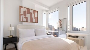 Ein modernes Hotelzimmer mit einem großen Bett, einem Nachttisch, einem Fenster mit Stadtblick und einer Wand-Klimaanlage.