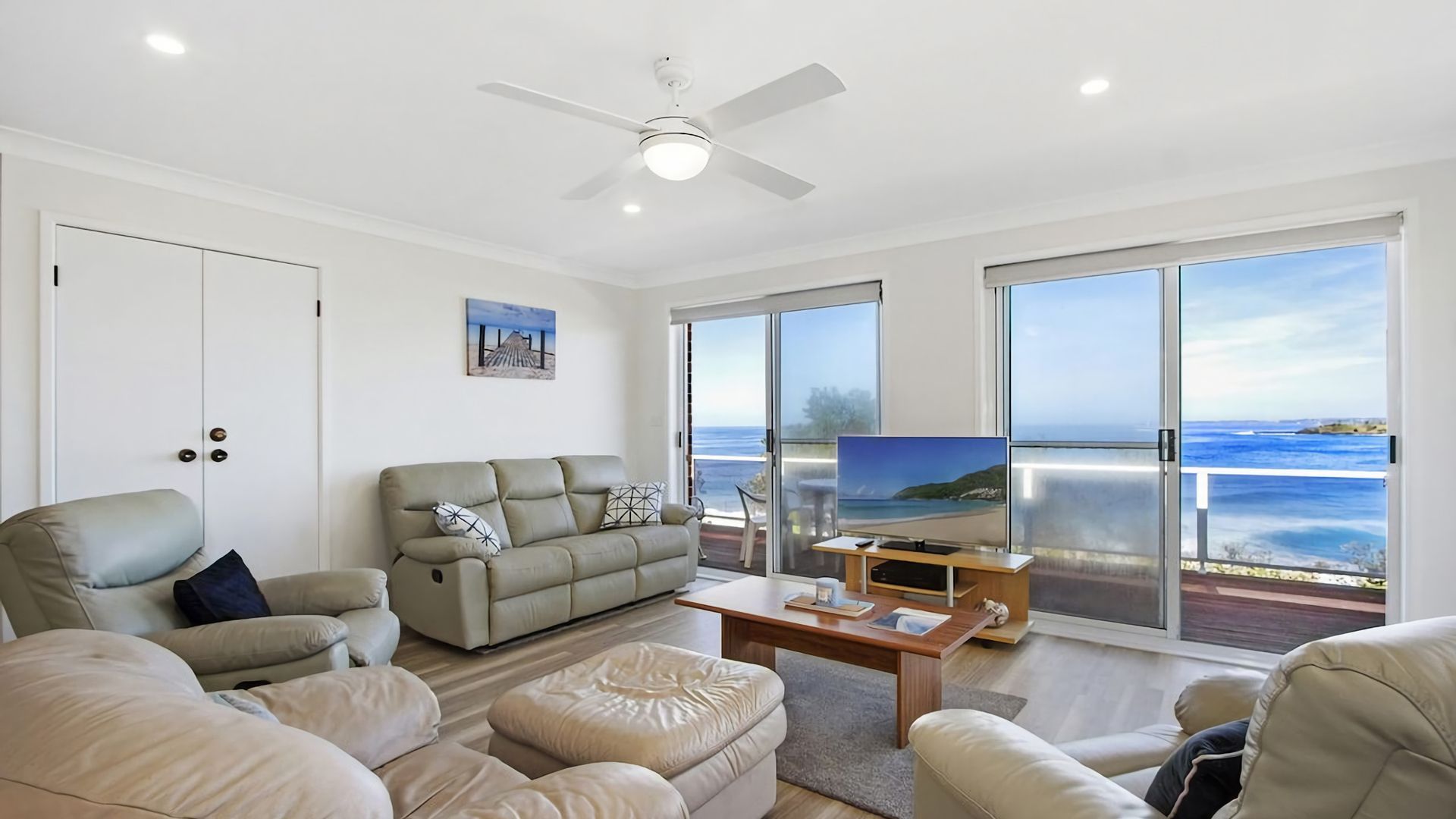 Una sala de estar moderna con sofá, sillones, una mesa de centro y televisor. Hay una puerta corrediza de vidrio que da a un balcón con vista al mar.
