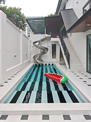 Ein moderner Außenpool mit einer Wendelrutsche, einem roten und grünen Schwimmspielzeug und einer Treppe zu einer erhöhten Ebene.