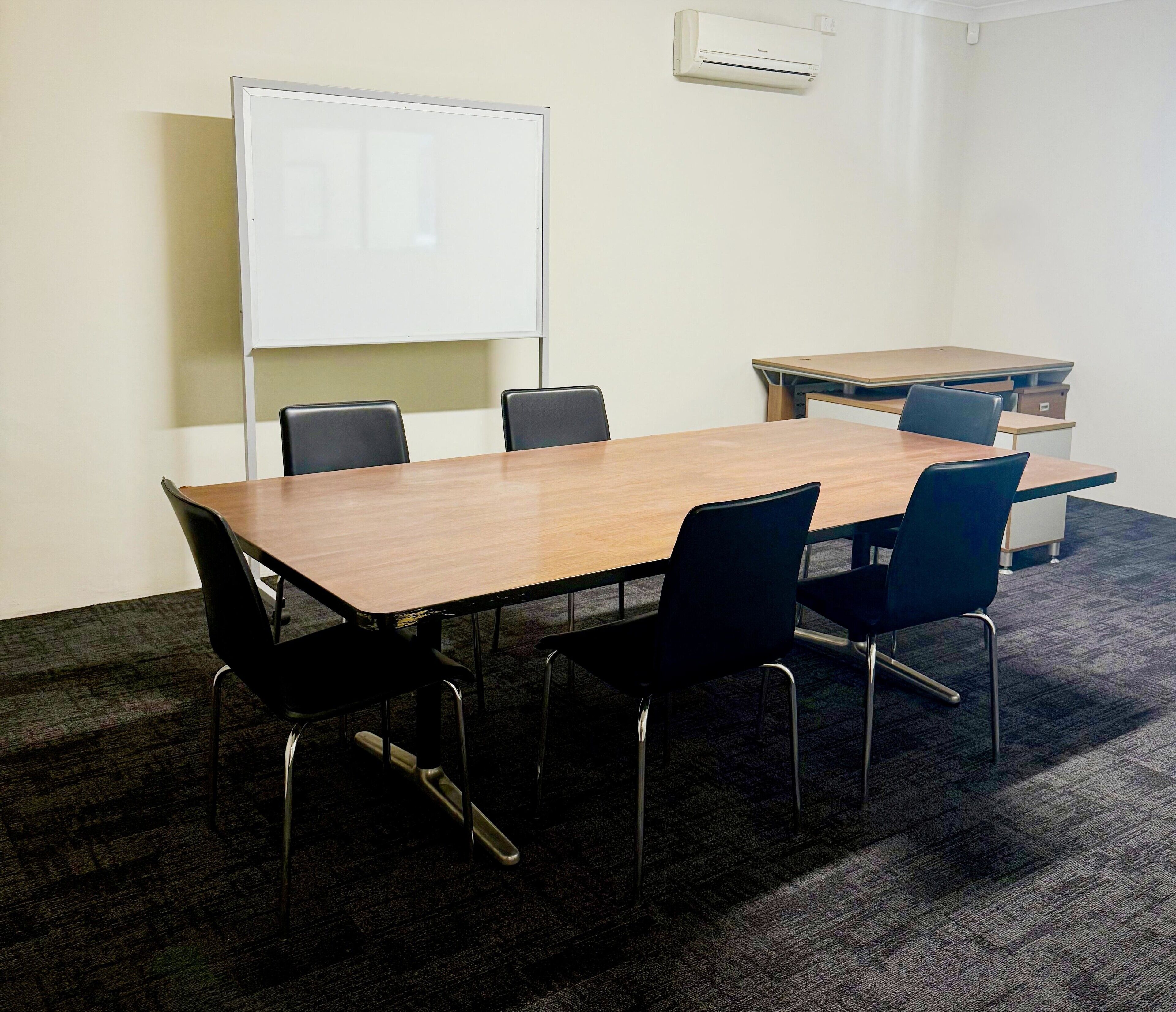 Una sala de conferencias con una mesa de madera, seis sillas negras, una pizarra y un escritorio pequeño con una computadora.