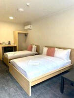 A hotel room with two beds, a desk, a chair, a coffee machine, and a wall-mounted air conditioner.