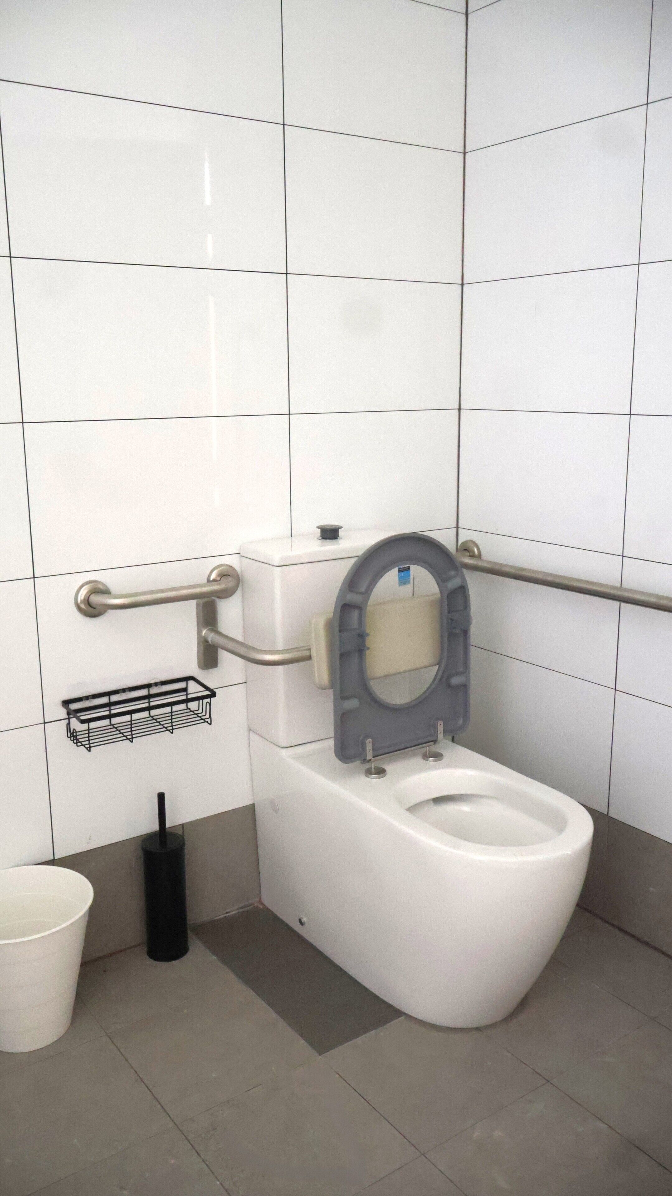 A modern bathroom with a white toilet, grab bars, a bidet, and a trash bin.