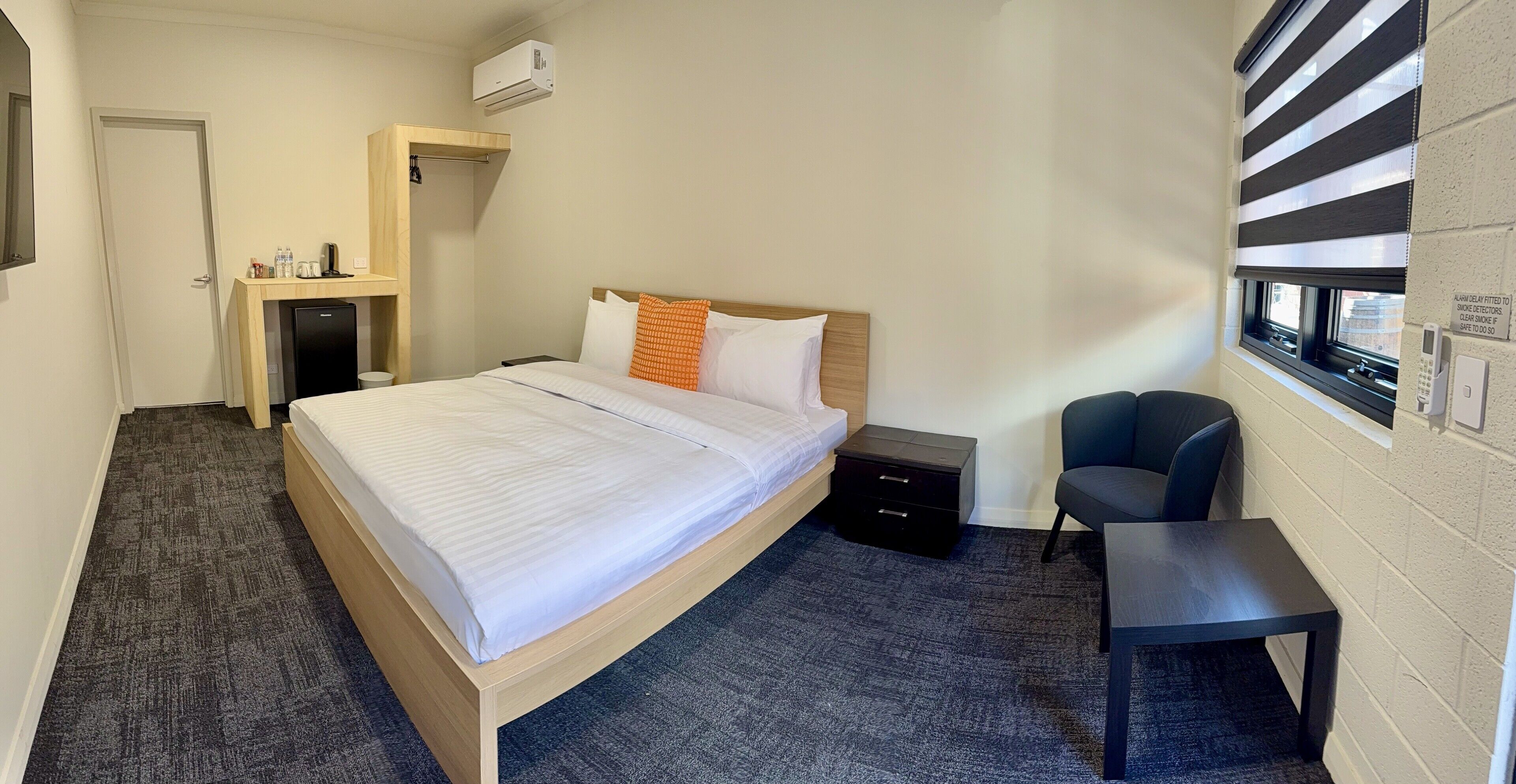A hotel room with a bed, a desk, a chair, a small refrigerator, and a window with blinds.