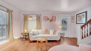 A cozy living room with a white sofa, a round wooden coffee table, and a pink ottoman. There is a small side table with a lamp, a window with curtains, and a door with a mirror. - The Allen House: Chic & Colorful 3BR Downtown Ellicottville (Ellicottville)