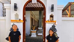 Deux personnes se tiennent devant l’entrée d’un bâtiment, qui est surmonté d’un toit recouvert de tuiles et dont la façade est blanche.