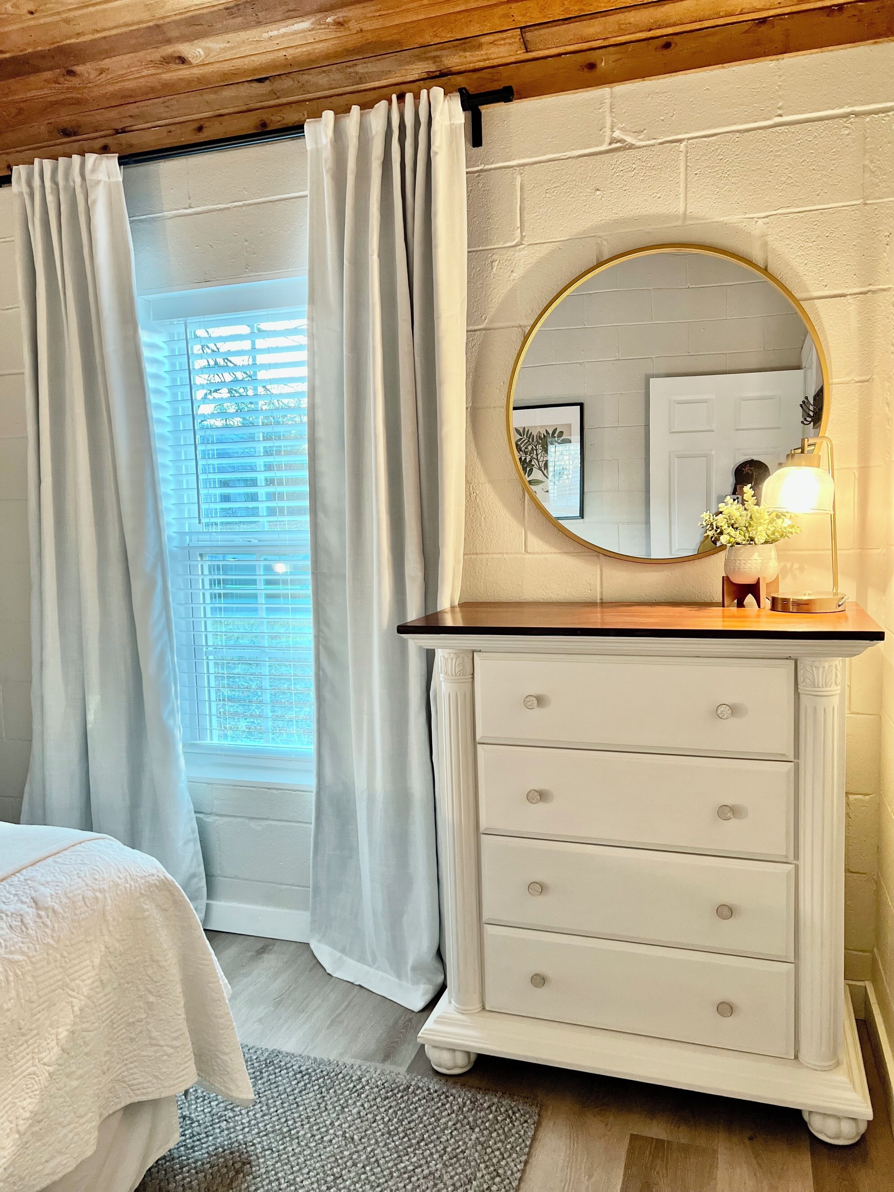 A bedroom with a bed, a dresser with drawers, a round mirror, and a window with curtains.