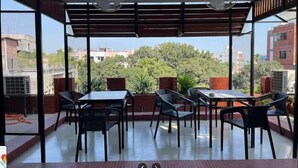 A covered outdoor seating area with tables and chairs, a view of buildings and trees, and a metal fence. - Hotel Riverview Kishorganj (Kishoreganj Sadar)
