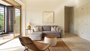 A modern living room with a sectional sofa, a round ottoman, and a wicker chair. There is a large window with blinds, a lamp on a side table, and a framed artwork on the wall.