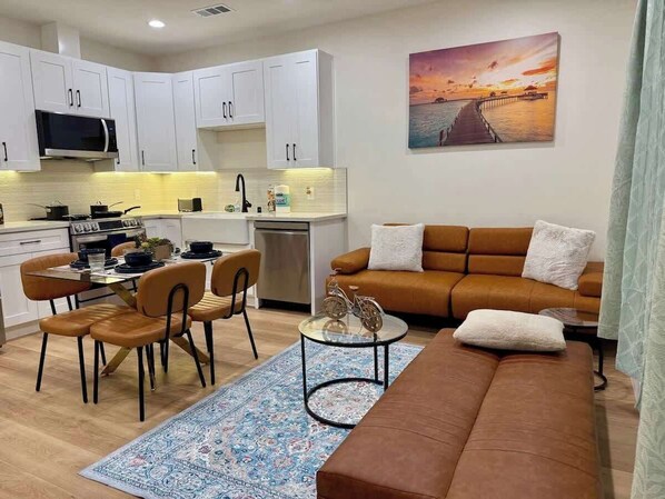 A modern kitchen with white cabinets, a dining area with a table and chairs, a sofa with cushions, and a painting of a sunset on the wall.