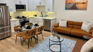 Une cuisine moderne avec des meubles blancs, un coin repas comprenant une table et des chaises, un canapé et un tableau représentant un coucher de soleil sur l’eau.
