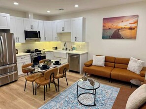 A modern kitchen with white cabinets, a dining area with a table and chairs, a sofa, and a painting of a sunset over water.