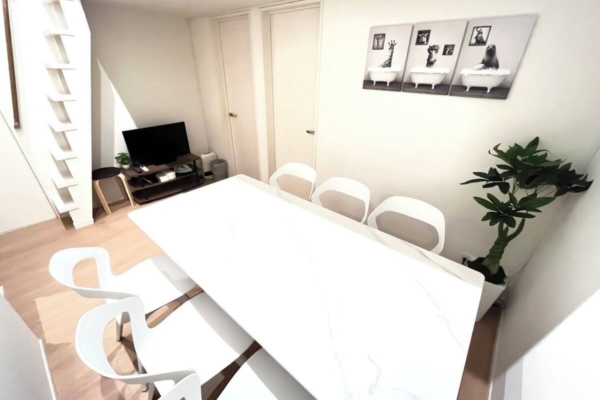 Een moderne eetruimte met een lange tafel en witte stoelen, een televisie en ingelijste foto's aan de muur.
