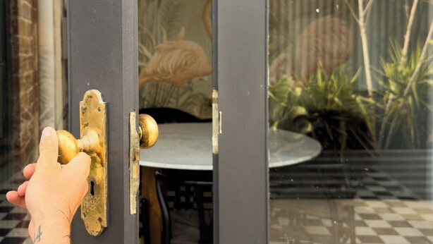 A door with a brass handle, a checkered floor, and a view of a garden with plants and a fence. - Forrest House Victor Harbor (Victor Harbor)