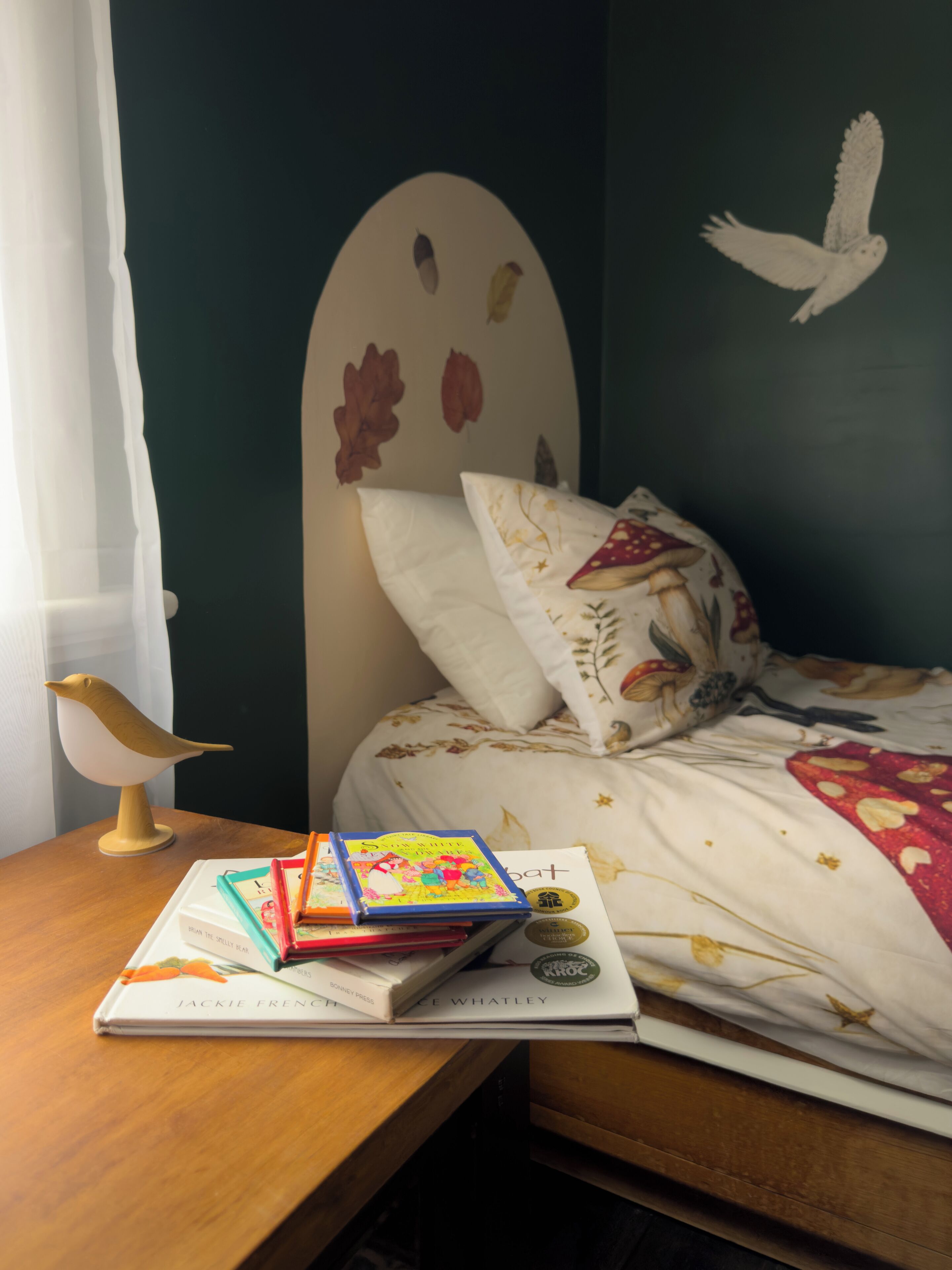 Un lit bien fait, avec une literie à motifs floraux, un mur orné d’une fresque représentant des oiseaux, et une table de chevet avec des livres et une lampe en forme d’oiseau.