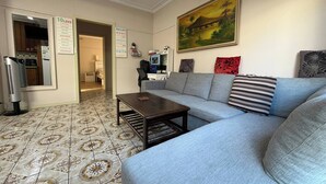 Un salon avec un canapé gris, une table basse, un tableau accroché au mur et un coin cuisine visible à travers une porte ouverte.