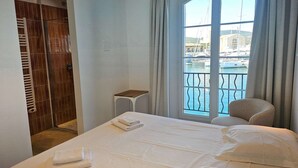 Une chambre d’hôtel comprenant un lit, une petite table, une chaise, une fenêtre donnant sur un port de plaisance et un dressing.