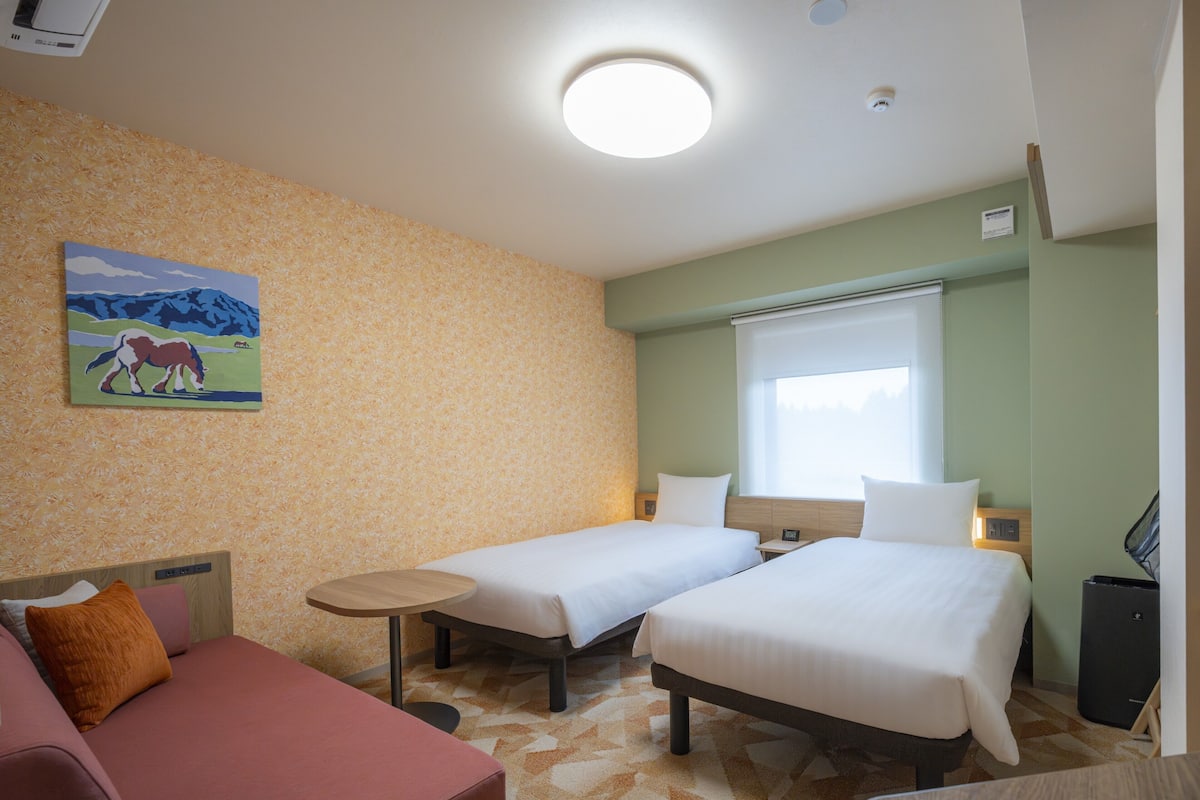 Habitación de hotel con dos camas, una mesita, un sofá, un cuadro en la pared y una ventana con persianas.